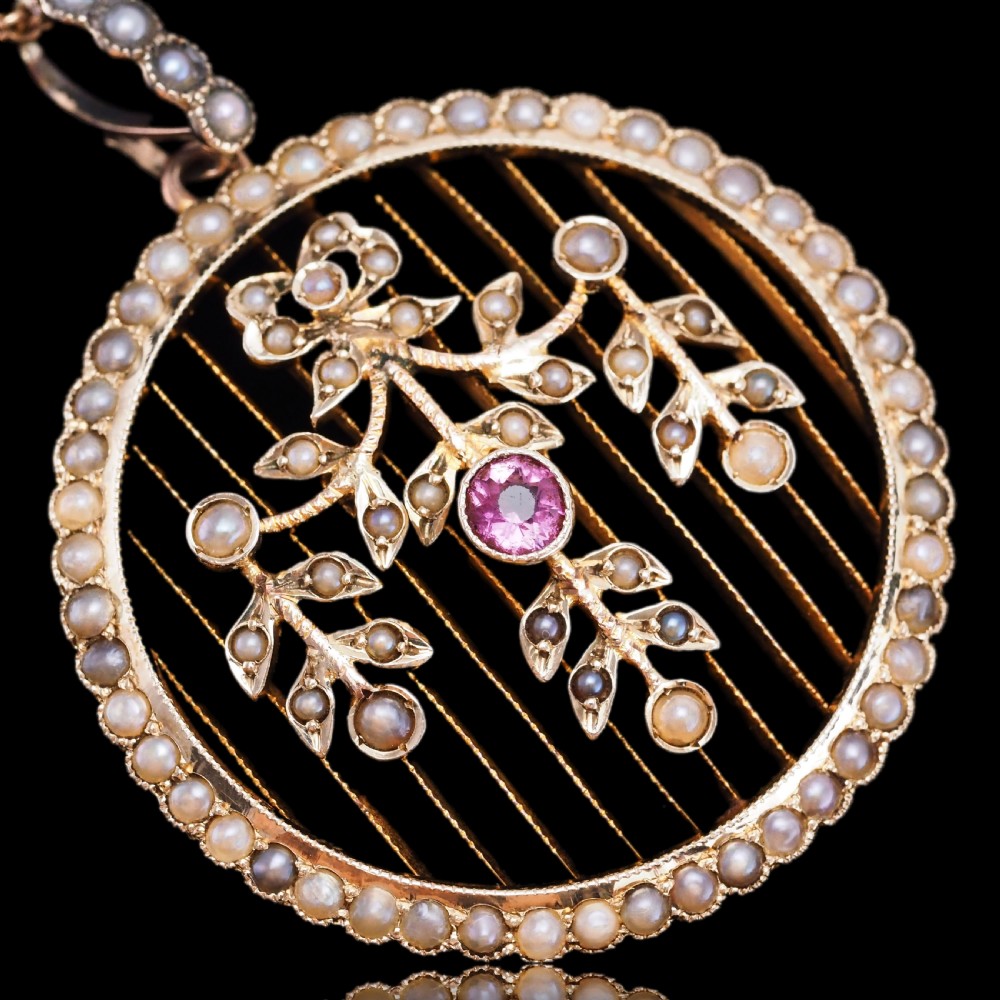 An Edwardian circular gold pendant featuring a halo of seed pearls framing an inner wire grid with a pearl-set floral garland and a central round pink tourmaline. View XVI