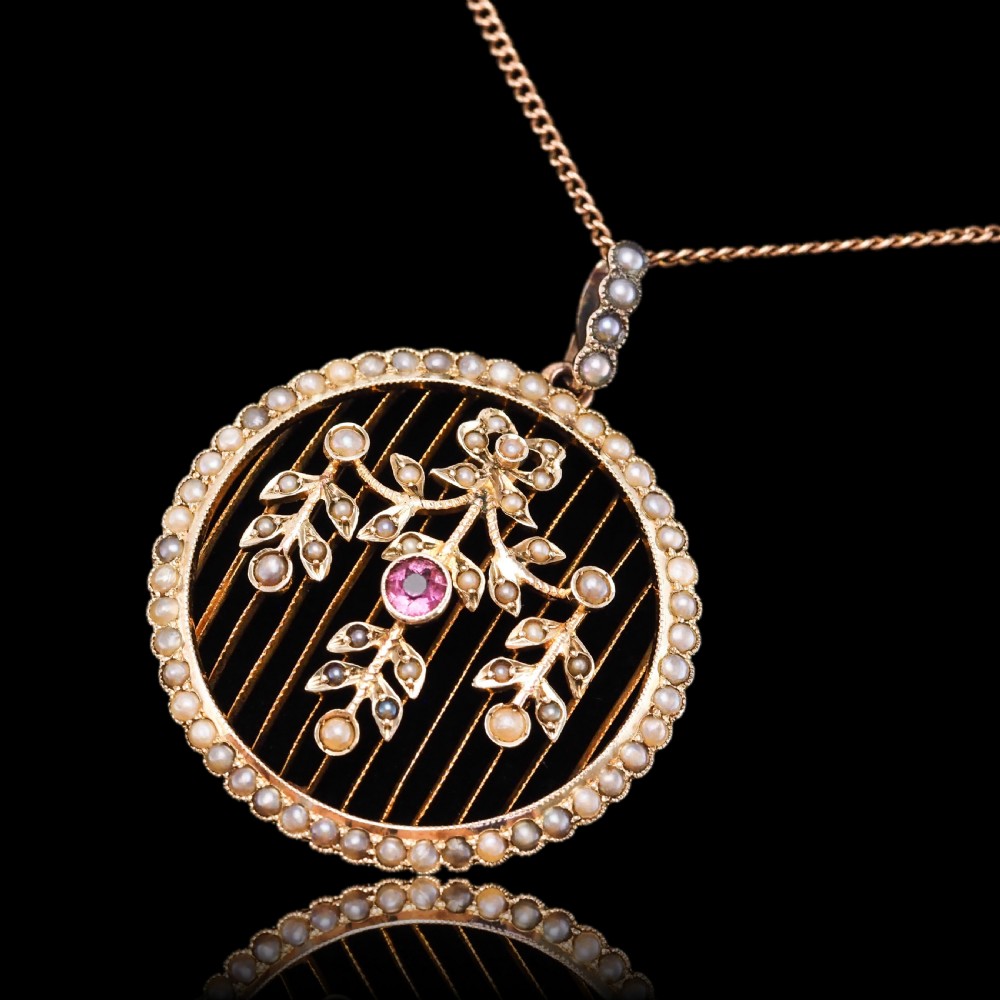 An Edwardian circular gold pendant featuring a halo of seed pearls framing an inner wire grid with a pearl-set floral garland and a central round pink tourmaline. View XVII