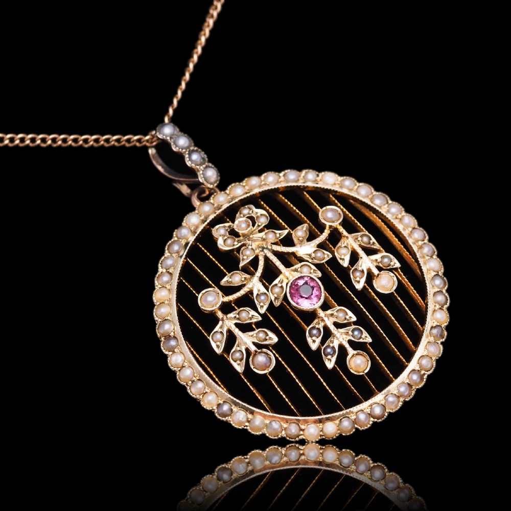An Edwardian circular gold pendant featuring a halo of seed pearls framing an inner wire grid with a pearl-set floral garland and a central round pink tourmaline. View XVIII