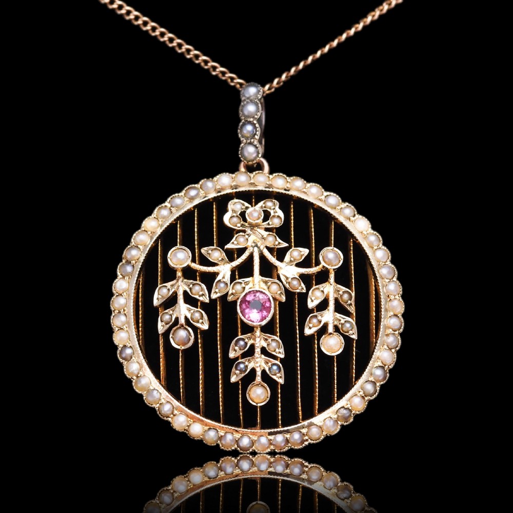 An Edwardian circular gold pendant featuring a halo of seed pearls framing an inner wire grid with a pearl-set floral garland and a central round pink tourmaline. View XIX