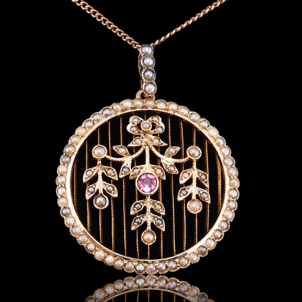 An Edwardian circular gold pendant featuring a halo of seed pearls framing an inner wire grid with a pearl-set floral garland and a central round pink tourmaline. View XX