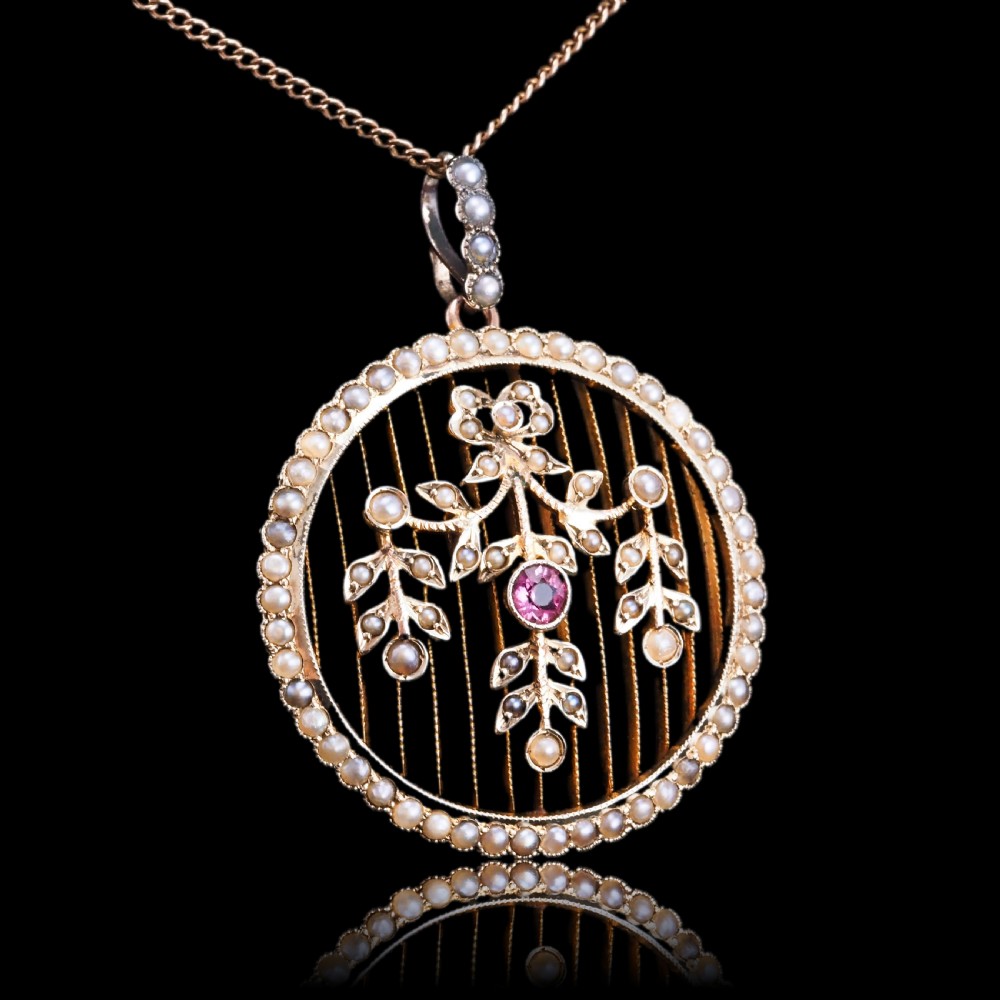 An Edwardian circular gold pendant featuring a halo of seed pearls framing an inner wire grid with a pearl-set floral garland and a central round pink tourmaline. View XXI