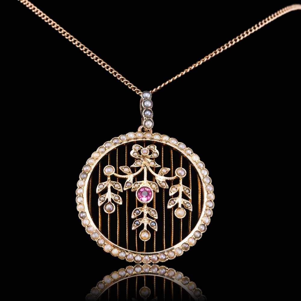 An Edwardian circular gold pendant featuring a halo of seed pearls framing an inner wire grid with a pearl-set floral garland and a central round pink tourmaline. View XXII