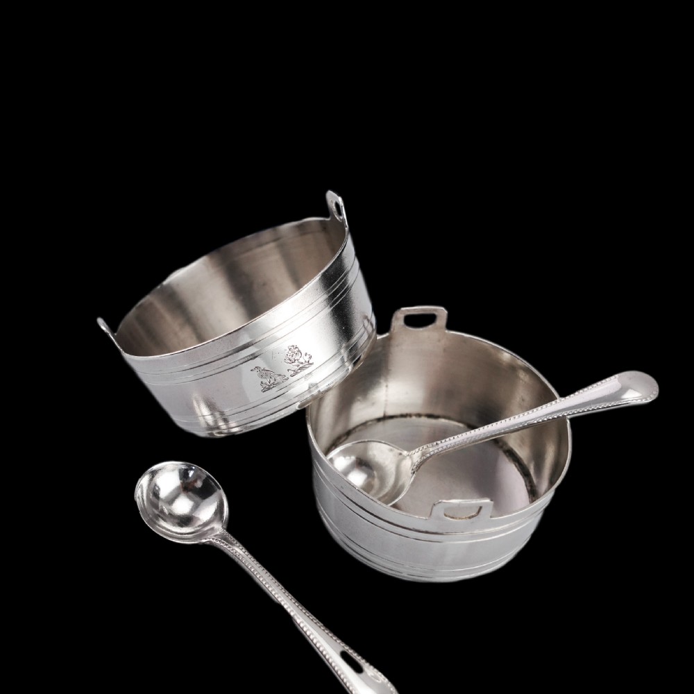 A pair of Victorian solid silver salt cellars crafted in the form of small pails with handles and engraved bird motifs, shown alongside two matching silver spoons. View X