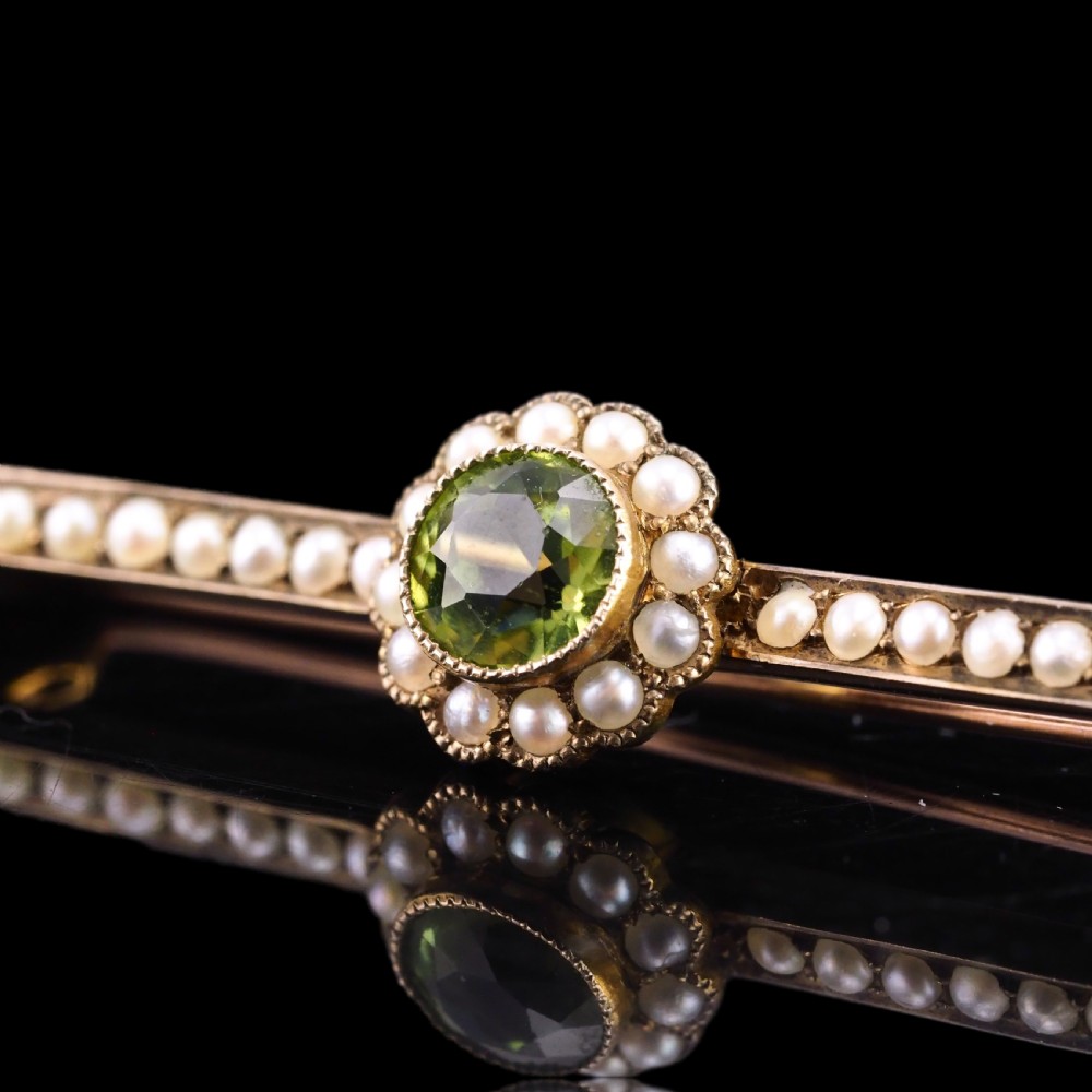 A 9ct gold late Victorian bar brooch featuring a central round faceted green peridot framed by a seed pearl halo, extending into pearl-set horizontal bars. View V