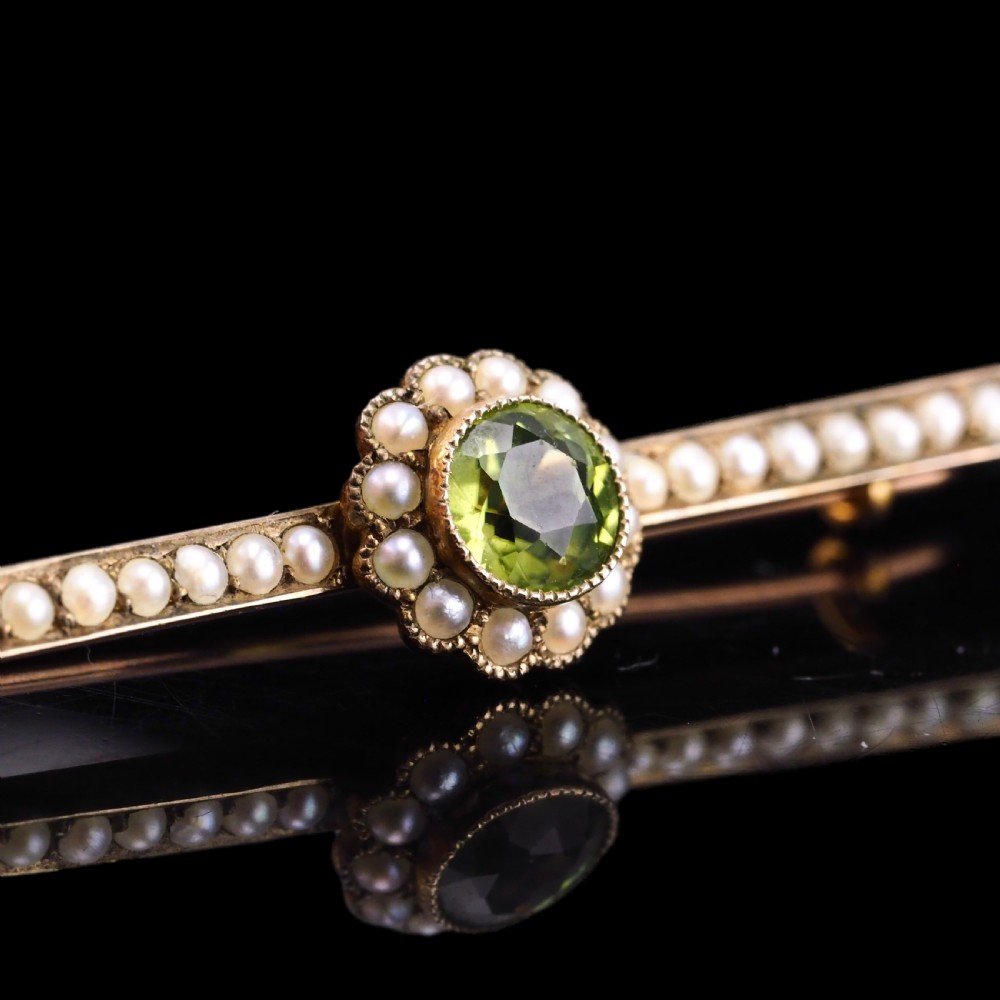 A 9ct gold late Victorian bar brooch featuring a central round faceted green peridot framed by a seed pearl halo, extending into pearl-set horizontal bars. View VI