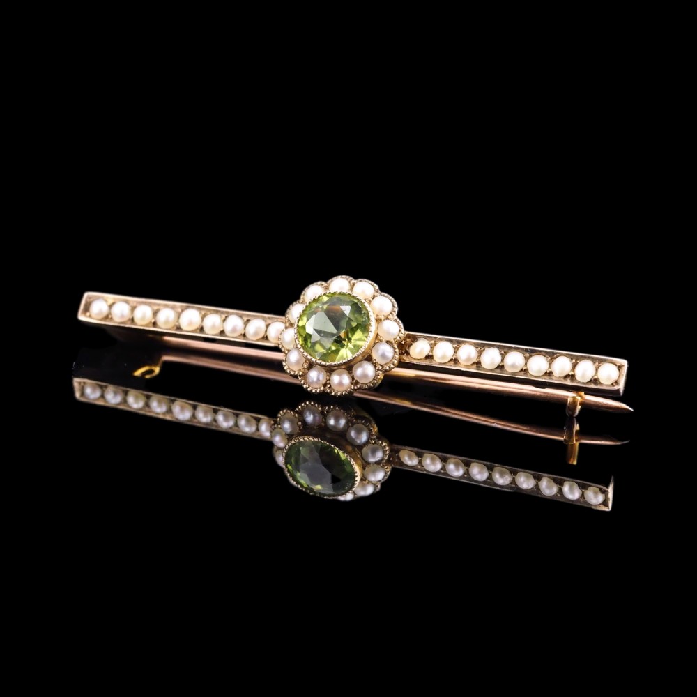 A 9ct gold late Victorian bar brooch featuring a central round faceted green peridot framed by a seed pearl halo, extending into pearl-set horizontal bars. View VII