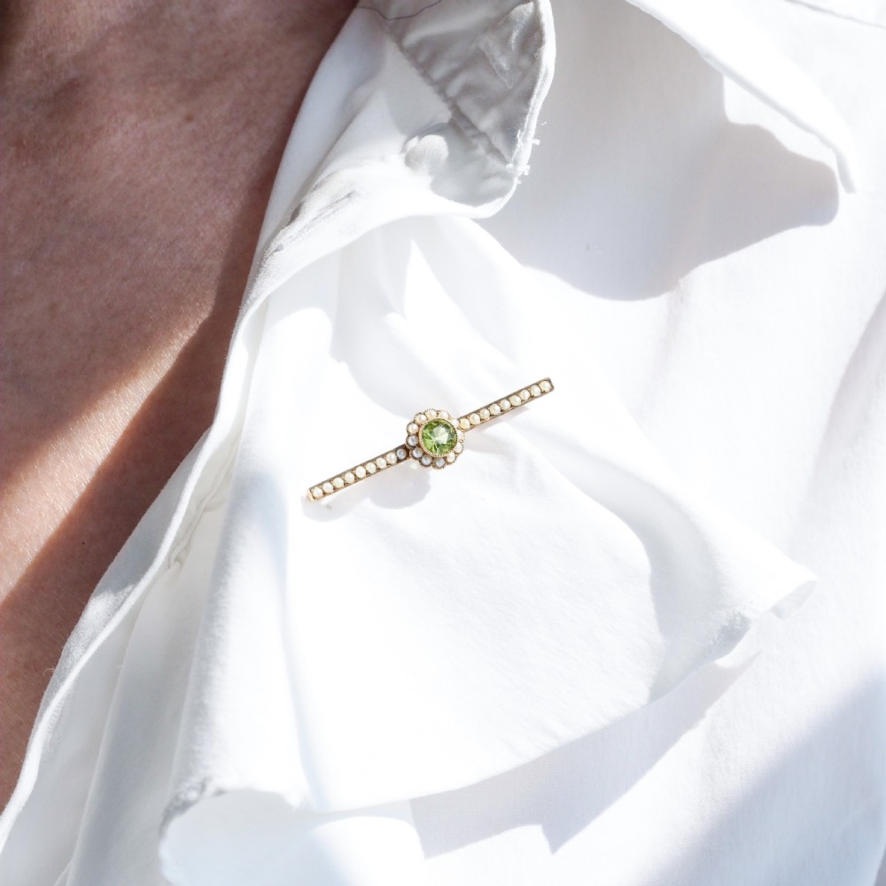 A 9ct gold late Victorian bar brooch featuring a central round faceted green peridot framed by a seed pearl halo, extending into pearl-set horizontal bars. View IX