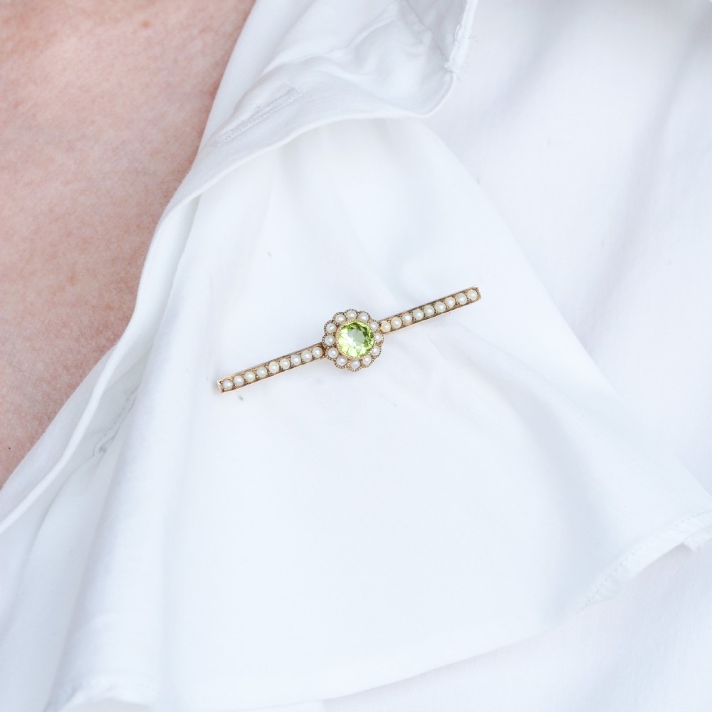 A 9ct gold late Victorian bar brooch featuring a central round faceted green peridot framed by a seed pearl halo, extending into pearl-set horizontal bars. View X