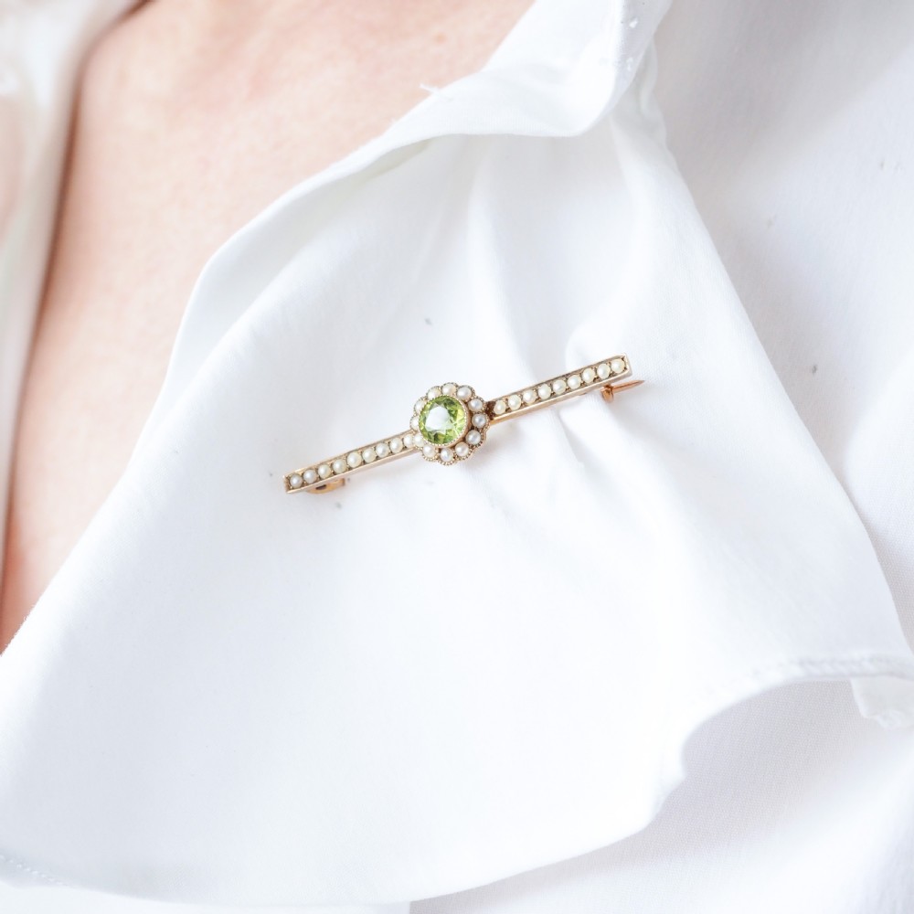 A 9ct gold late Victorian bar brooch featuring a central round faceted green peridot framed by a seed pearl halo, extending into pearl-set horizontal bars. View XII