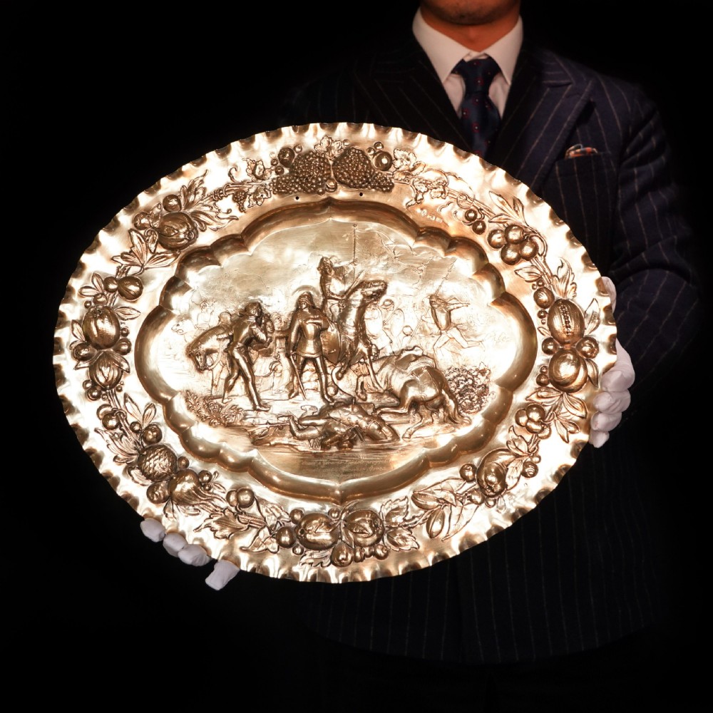 An oval 19th-century solid silver gilt charger dish featuring a central raised relief of a battle scene surrounded by embossed fruit borders. View II