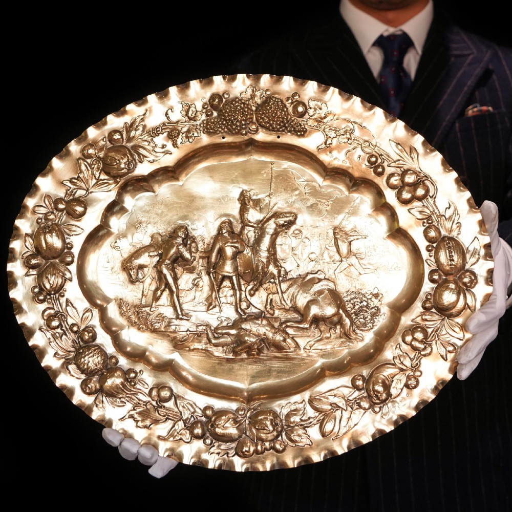 An oval 19th-century solid silver gilt charger dish featuring a central raised relief of a battle scene surrounded by embossed fruit borders. View V
