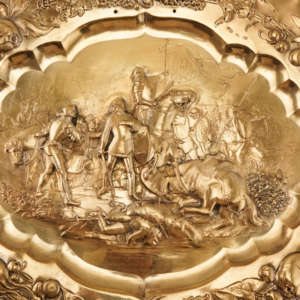 An oval 19th-century solid silver gilt charger dish featuring a central raised relief of a battle scene surrounded by embossed fruit borders. View X