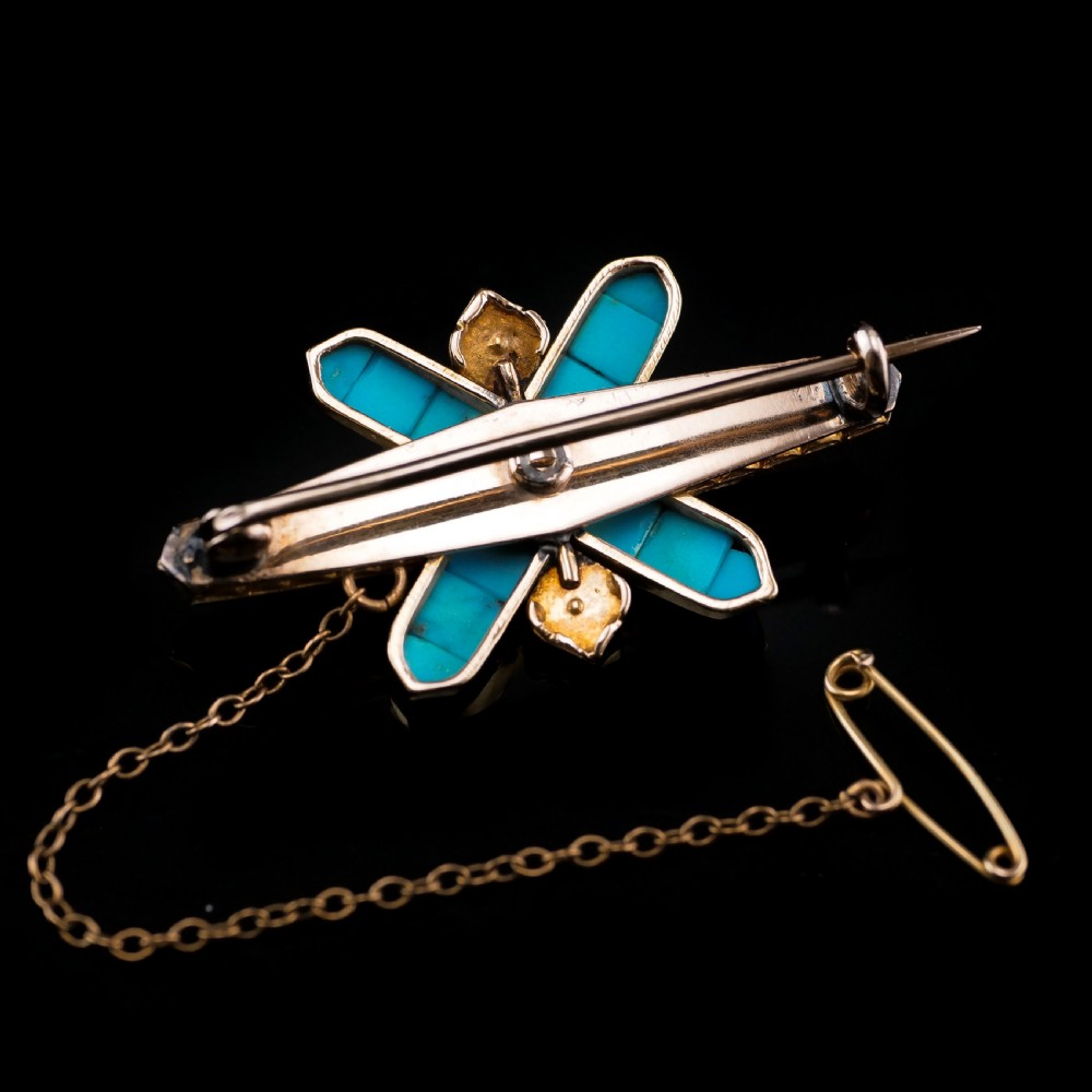 An antique Victorian gold cross-shaped brooch set with cabochon turquoise, white seed pearls, a central diamond, and attached to a safety chain. View X