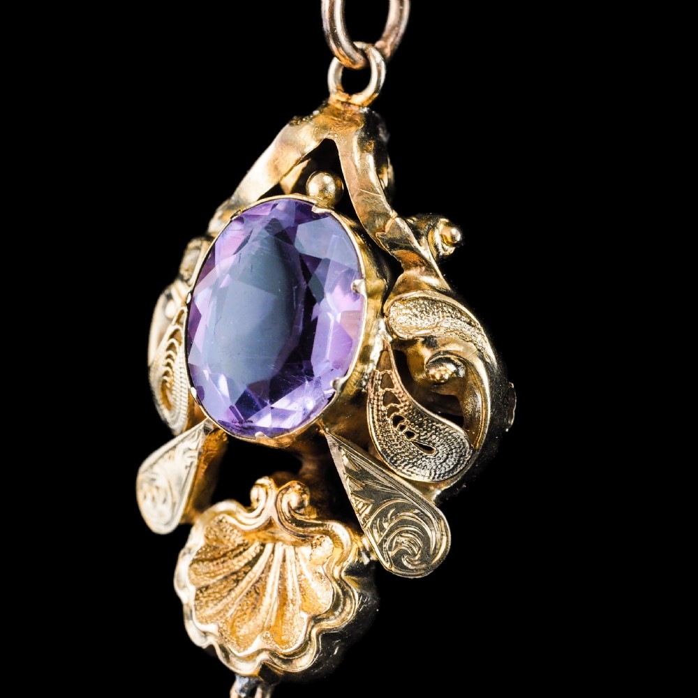 An antique 14ct yellow gold Victorian pendant featuring a central oval faceted amethyst surrounded by scrollwork, a shell motif, and an articulated drop element. View XVI