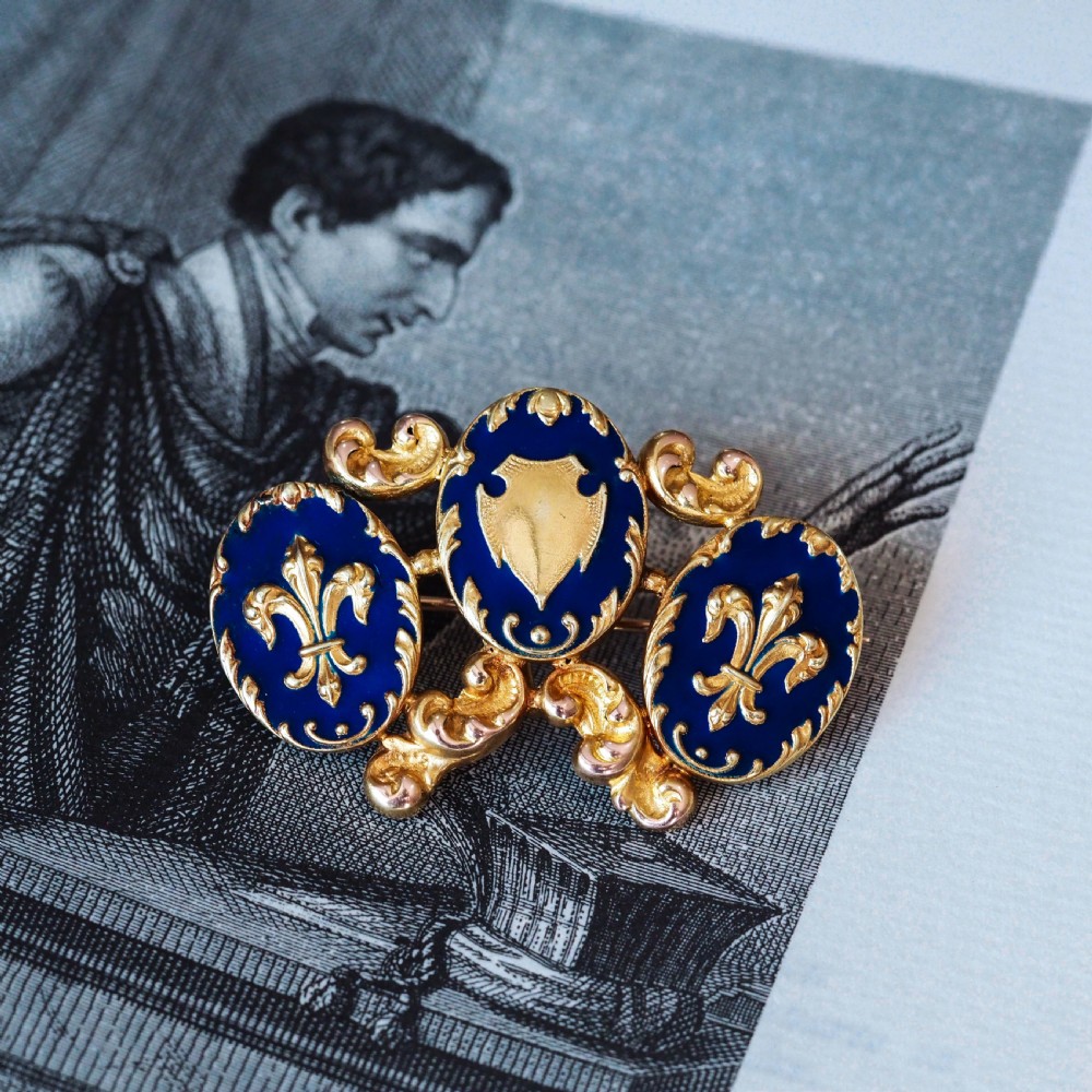 A 19th-century 18ct gold brooch featuring three dark blue enamel oval cartouches decorated with gold heraldic motifs and rococo scrollwork. View V