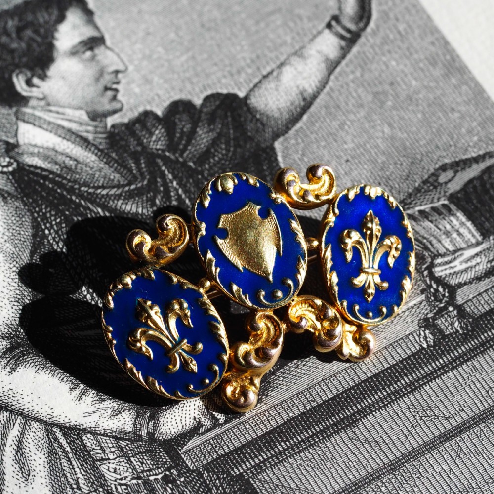 A 19th-century 18ct gold brooch featuring three dark blue enamel oval cartouches decorated with gold heraldic motifs and rococo scrollwork. View VI