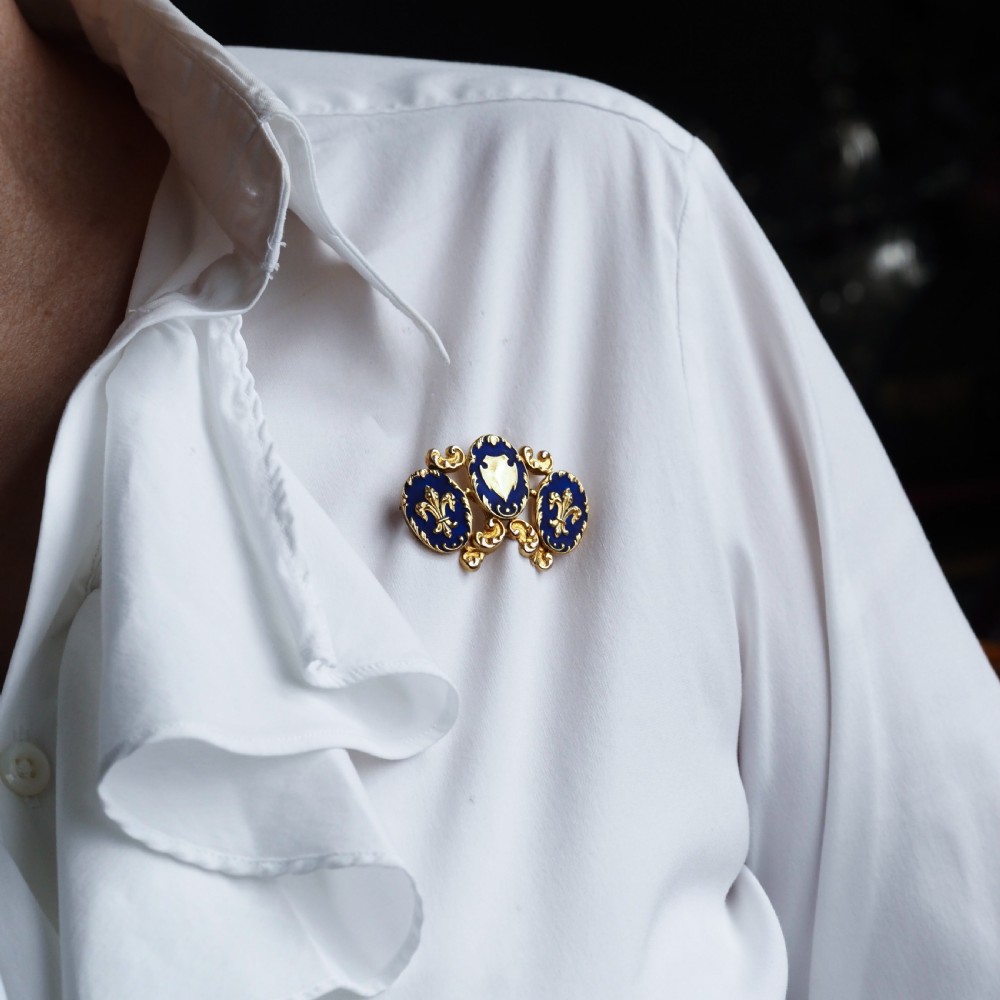 A 19th-century 18ct gold brooch featuring three dark blue enamel oval cartouches decorated with gold heraldic motifs and rococo scrollwork. View X