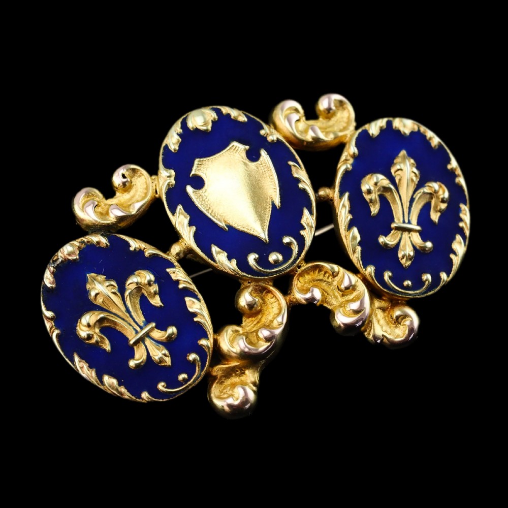 A 19th-century 18ct gold brooch featuring three dark blue enamel oval cartouches decorated with gold heraldic motifs and rococo scrollwork. View XVIII