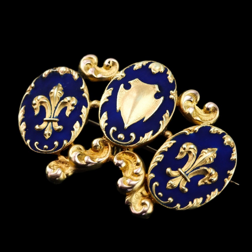 A 19th-century 18ct gold brooch featuring three dark blue enamel oval cartouches decorated with gold heraldic motifs and rococo scrollwork. View XIX