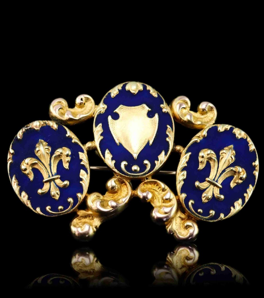 A 19th-century 18ct gold brooch featuring three dark blue enamel oval cartouches decorated with gold heraldic motifs and rococo scrollwork.