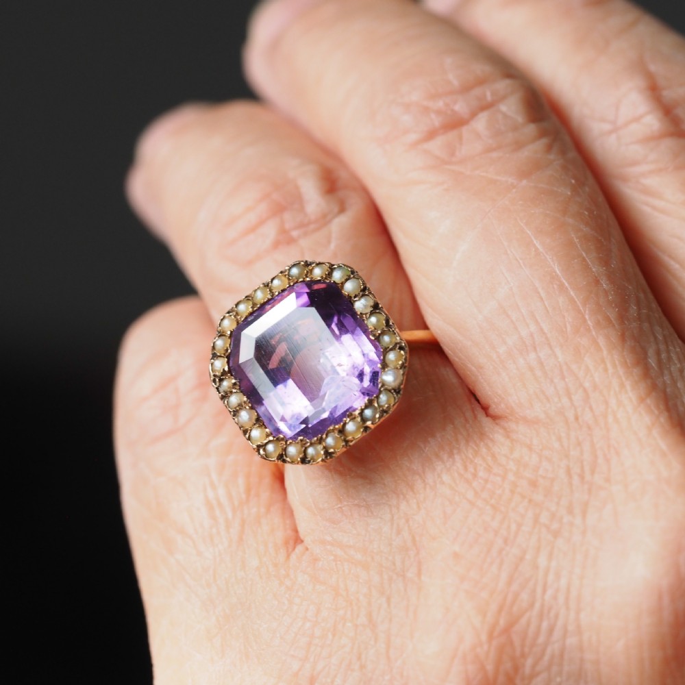 An Edwardian 18ct gold ring featuring a central emerald-cut purple amethyst surrounded by a halo of small seed pearls.