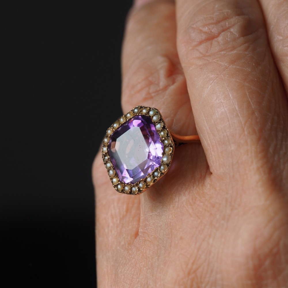 An Edwardian 18ct gold ring featuring a central emerald-cut purple amethyst surrounded by a halo of small seed pearls. View II