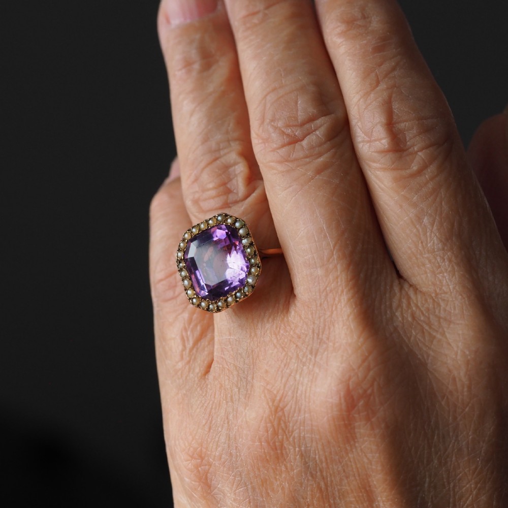 An Edwardian 18ct gold ring featuring a central emerald-cut purple amethyst surrounded by a halo of small seed pearls. View III