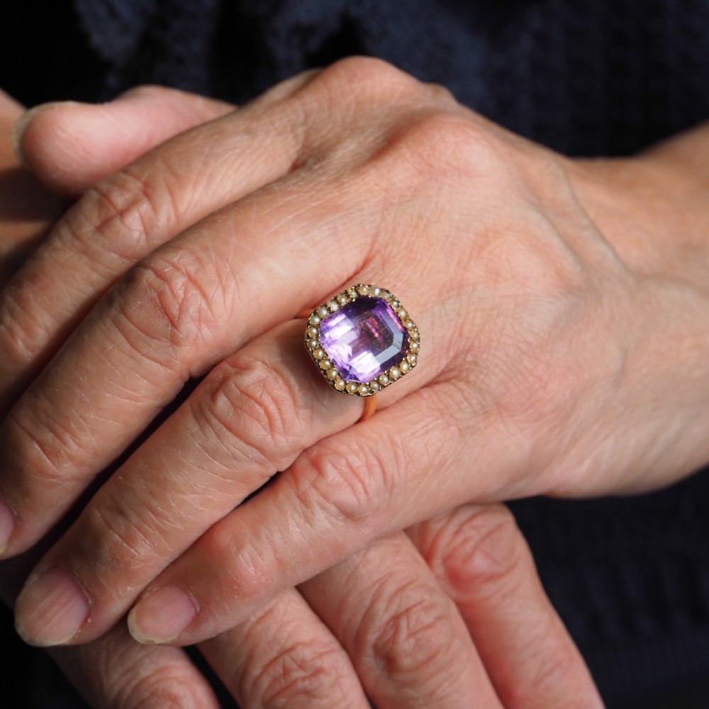 An Edwardian 18ct gold ring featuring a central emerald-cut purple amethyst surrounded by a halo of small seed pearls. View V
