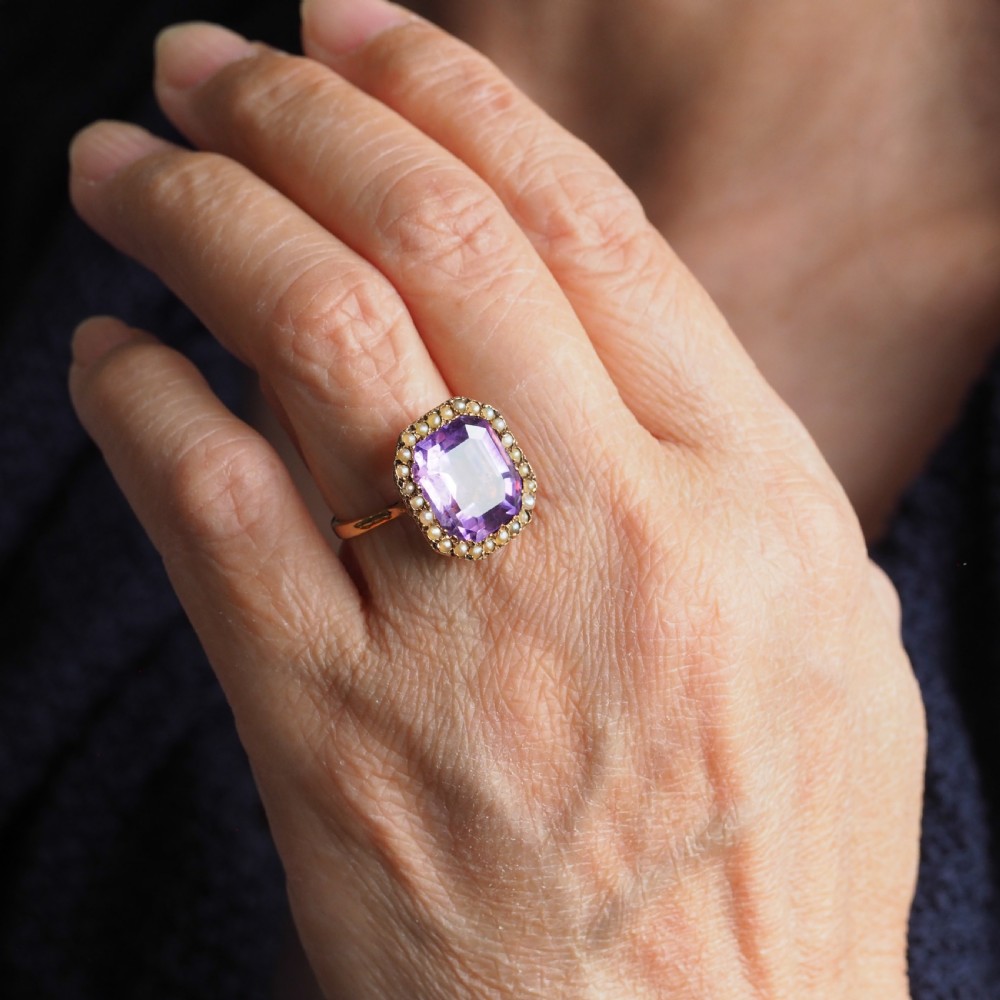 An Edwardian 18ct gold ring featuring a central emerald-cut purple amethyst surrounded by a halo of small seed pearls. View VI