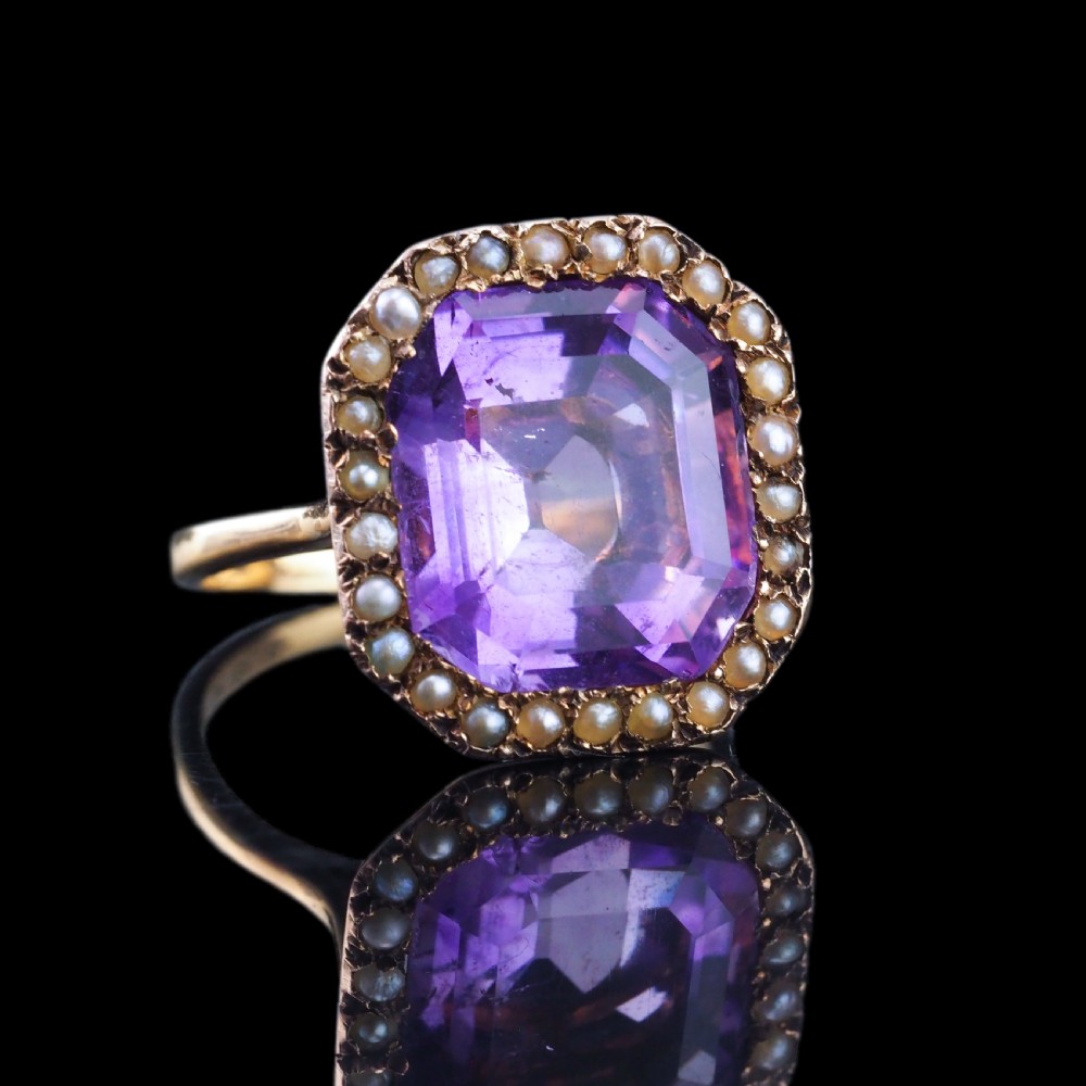 An Edwardian 18ct gold ring featuring a central emerald-cut purple amethyst surrounded by a halo of small seed pearls. View VII