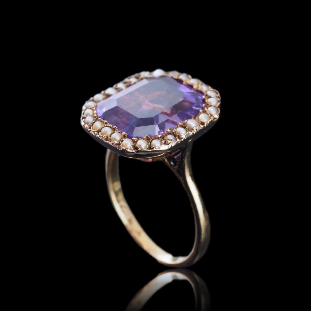 An Edwardian 18ct gold ring featuring a central emerald-cut purple amethyst surrounded by a halo of small seed pearls. View X