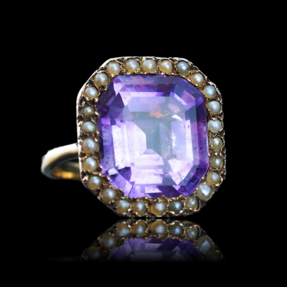 An Edwardian 18ct gold ring featuring a central emerald-cut purple amethyst surrounded by a halo of small seed pearls. View XV