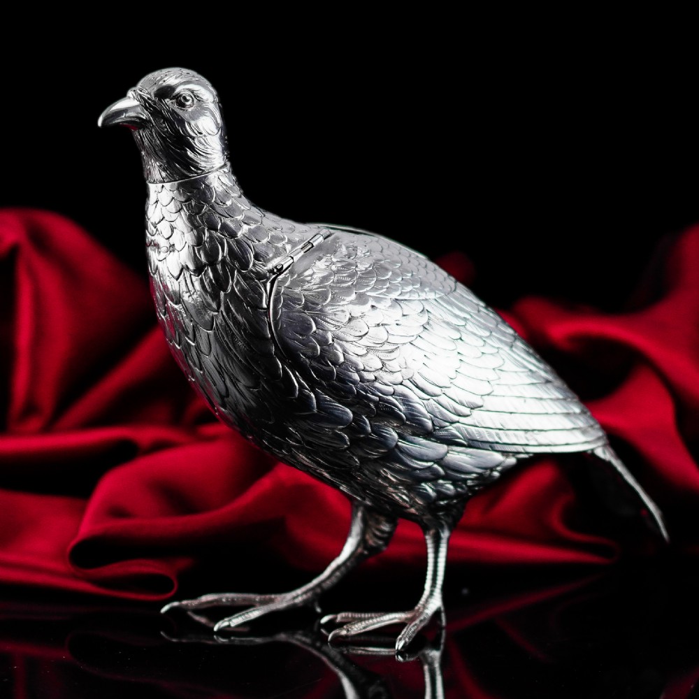 A solid silver model of a partridge with chased feather detailing standing on two feet with a hinged compartment lid along its back.