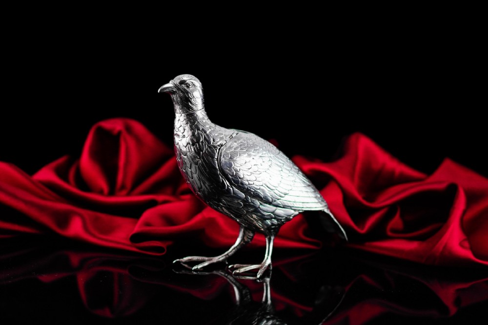A solid silver model of a partridge with chased feather detailing standing on two feet with a hinged compartment lid along its back. View II