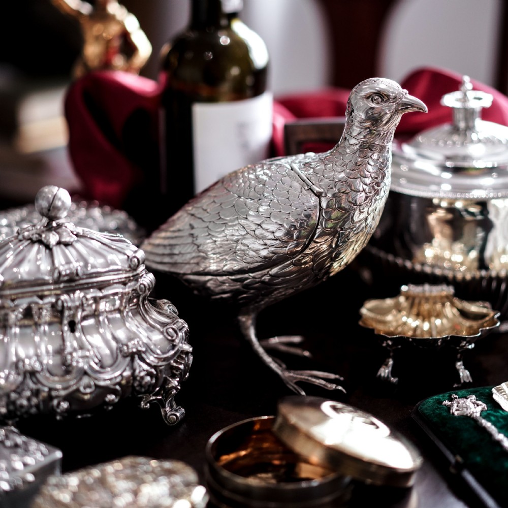 A solid silver model of a partridge with chased feather detailing standing on two feet with a hinged compartment lid along its back. View IX
