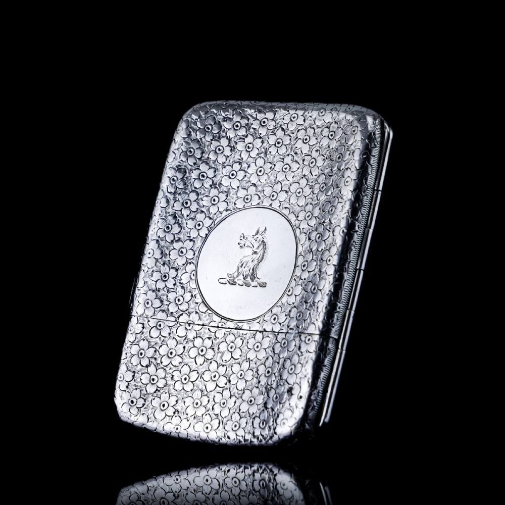 An antique Victorian sterling silver cigarette case decorated with an overall engraved floral pattern and a central oval cartouche containing a heraldic crest.