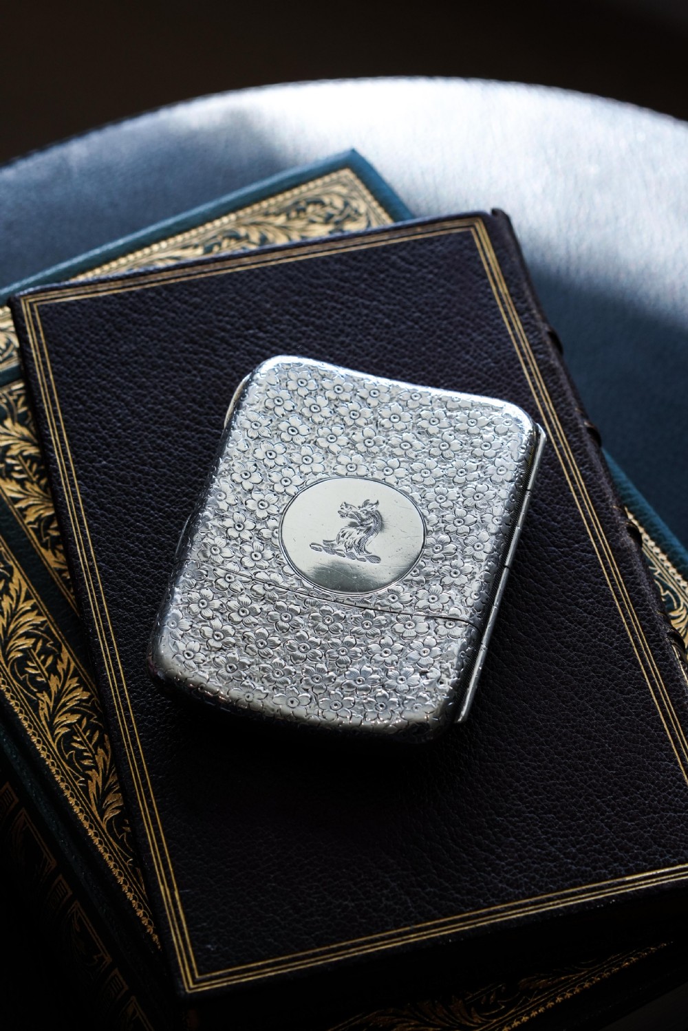 An antique Victorian sterling silver cigarette case decorated with an overall engraved floral pattern and a central oval cartouche containing a heraldic crest. View II
