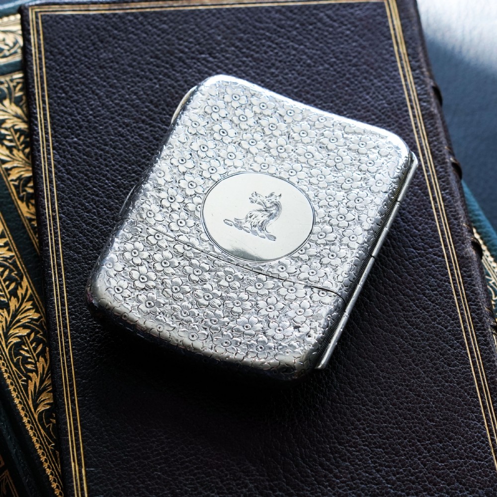 An antique Victorian sterling silver cigarette case decorated with an overall engraved floral pattern and a central oval cartouche containing a heraldic crest. View V