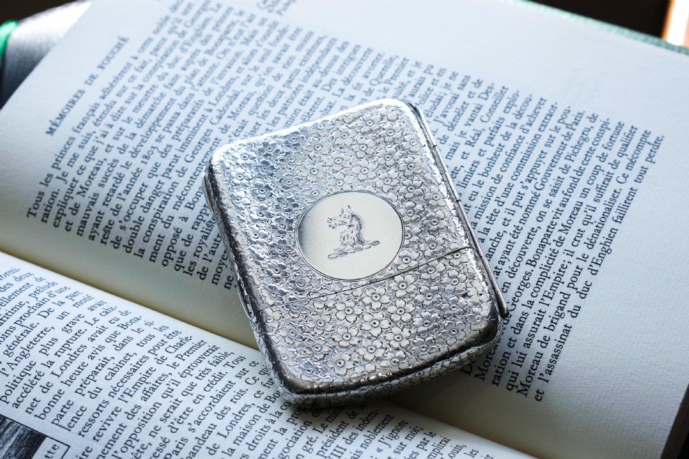 An antique Victorian sterling silver cigarette case decorated with an overall engraved floral pattern and a central oval cartouche containing a heraldic crest. View VI