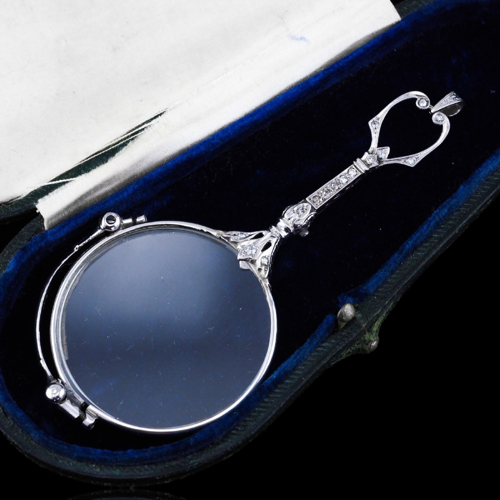 An Art Deco platinum lorgnette featuring a circular folding magnifying glass lens attached to a geometric handle set with small diamonds. View V