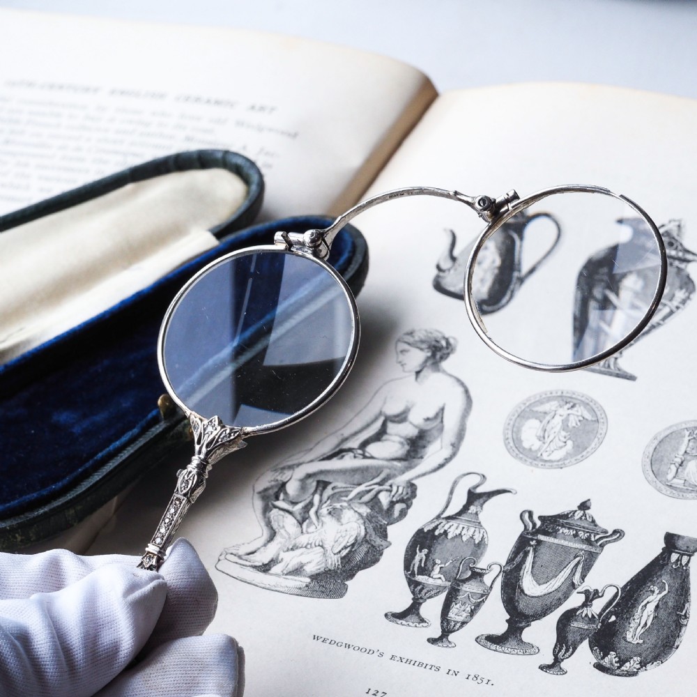 An Art Deco platinum lorgnette featuring a circular folding magnifying glass lens attached to a geometric handle set with small diamonds. View X