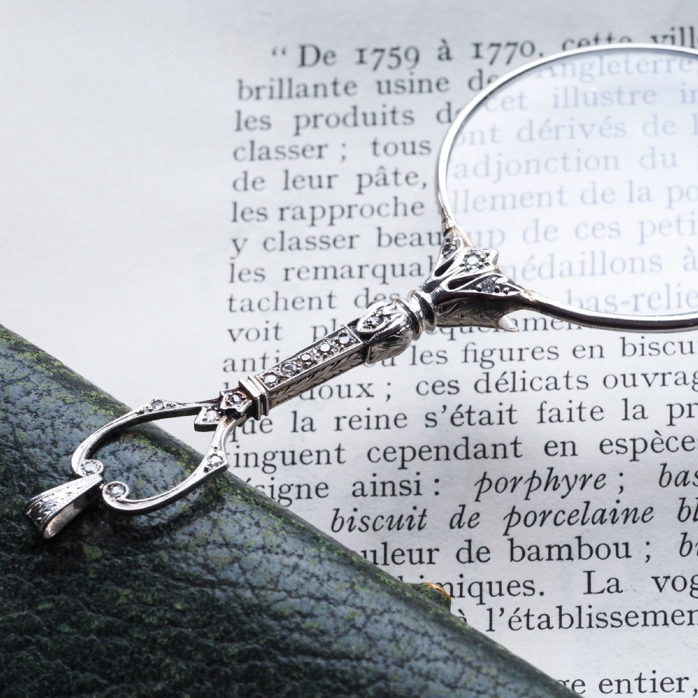 An Art Deco platinum lorgnette featuring a circular folding magnifying glass lens attached to a geometric handle set with small diamonds. View XVI