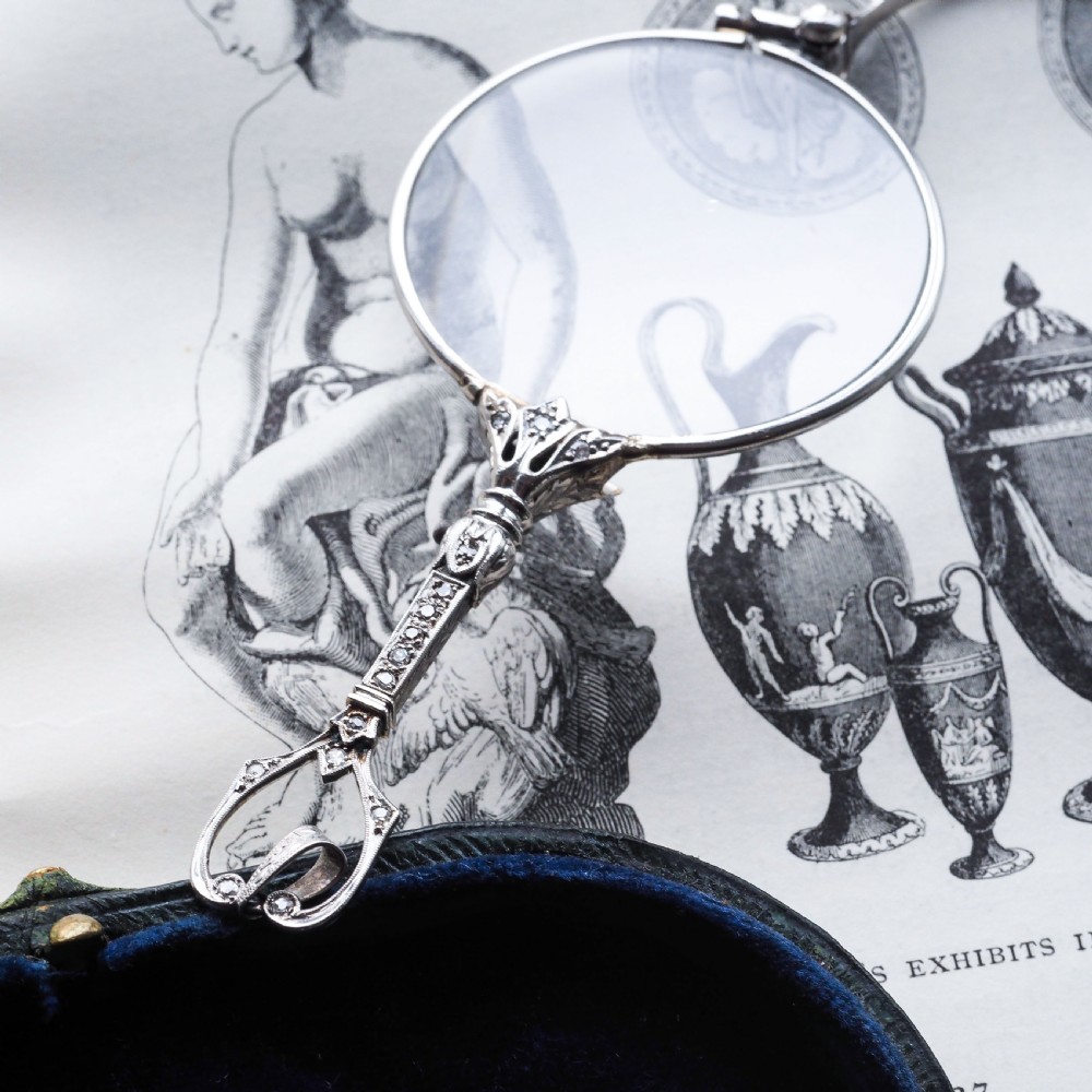 An Art Deco platinum lorgnette featuring a circular folding magnifying glass lens attached to a geometric handle set with small diamonds. View XVIII