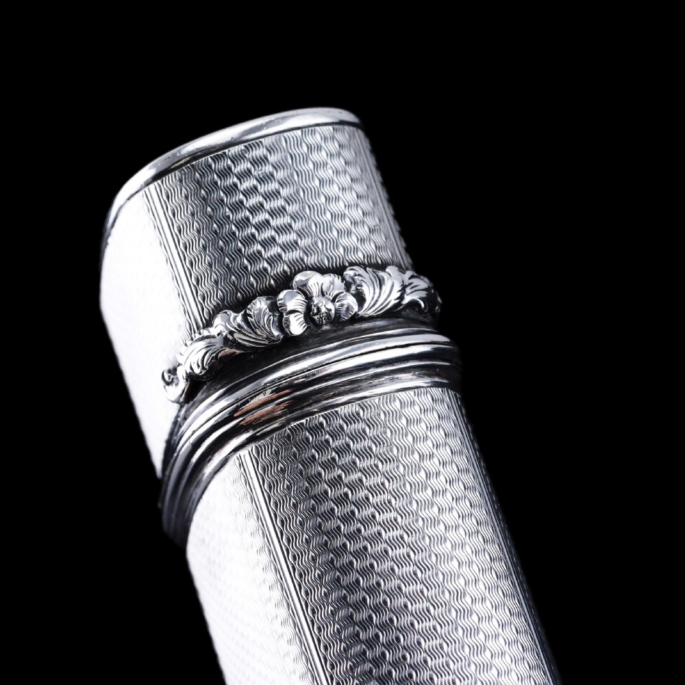 An antique engine-turned solid silver cigar case featuring floral relief thumbpiece detailing and an engraved cartouche on the end. View XVI