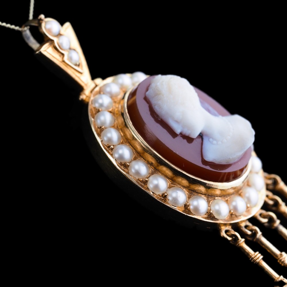 A Victorian 14K gold oval pendant featuring a carved white shell cameo profile against a brown background, framed by a border of seed pearls with articulated pearl drops. View X