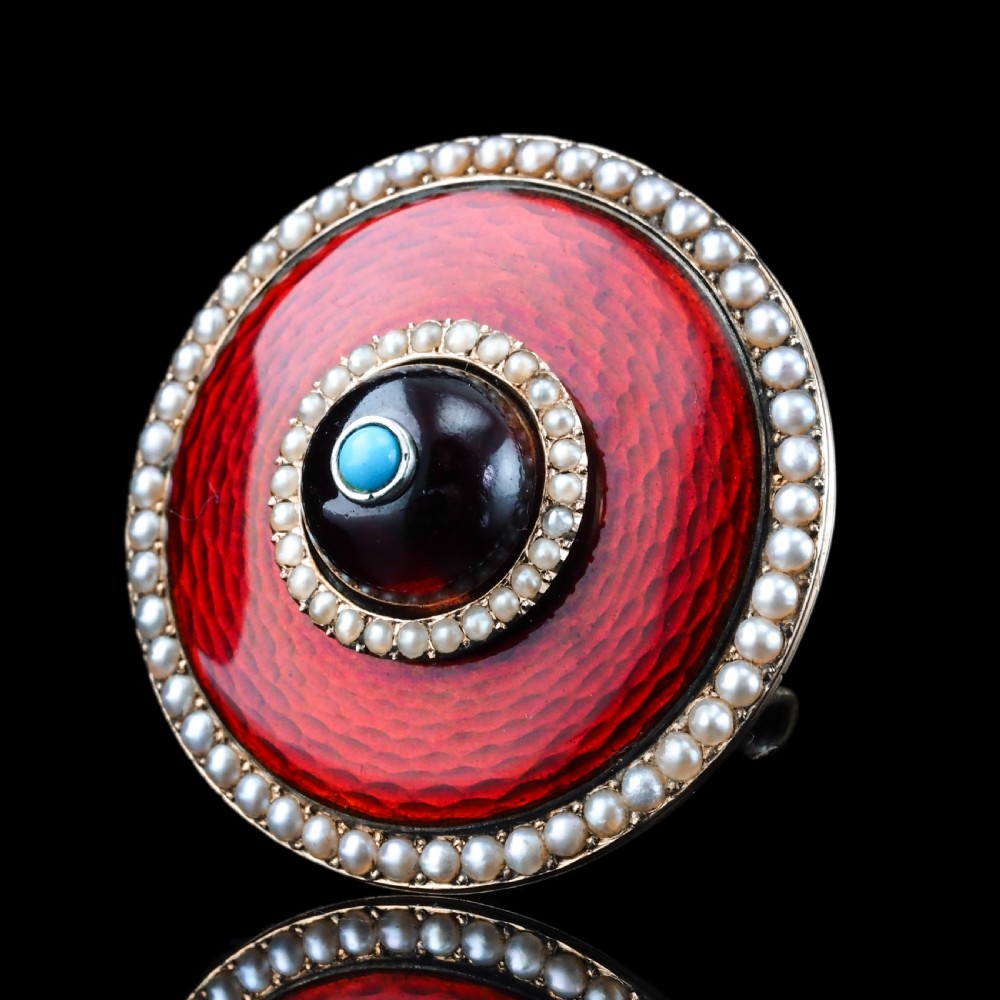 A circular Victorian red guilloche enamel brooch featuring concentric seed pearl borders, a central dark garnet cabochon, and a small turquoise accent set in gold.