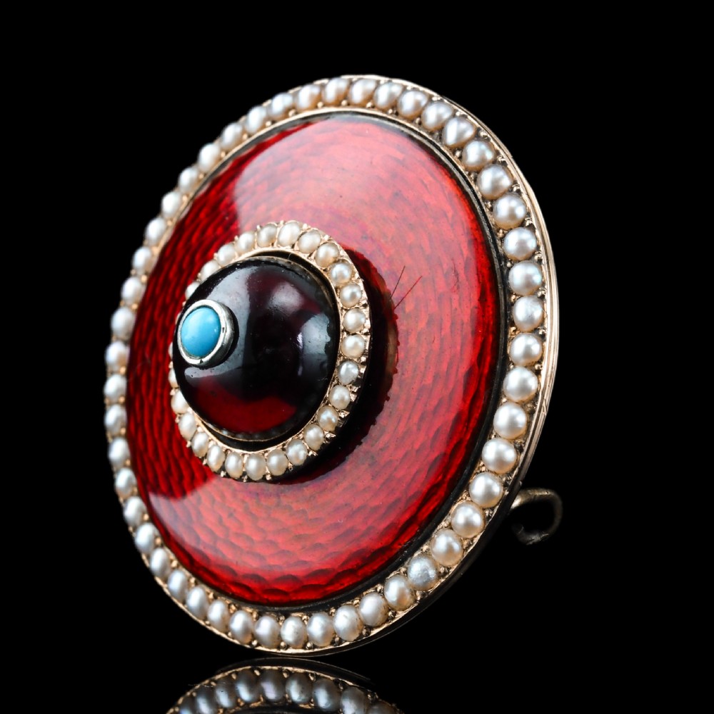 A circular Victorian red guilloche enamel brooch featuring concentric seed pearl borders, a central dark garnet cabochon, and a small turquoise accent set in gold. View II