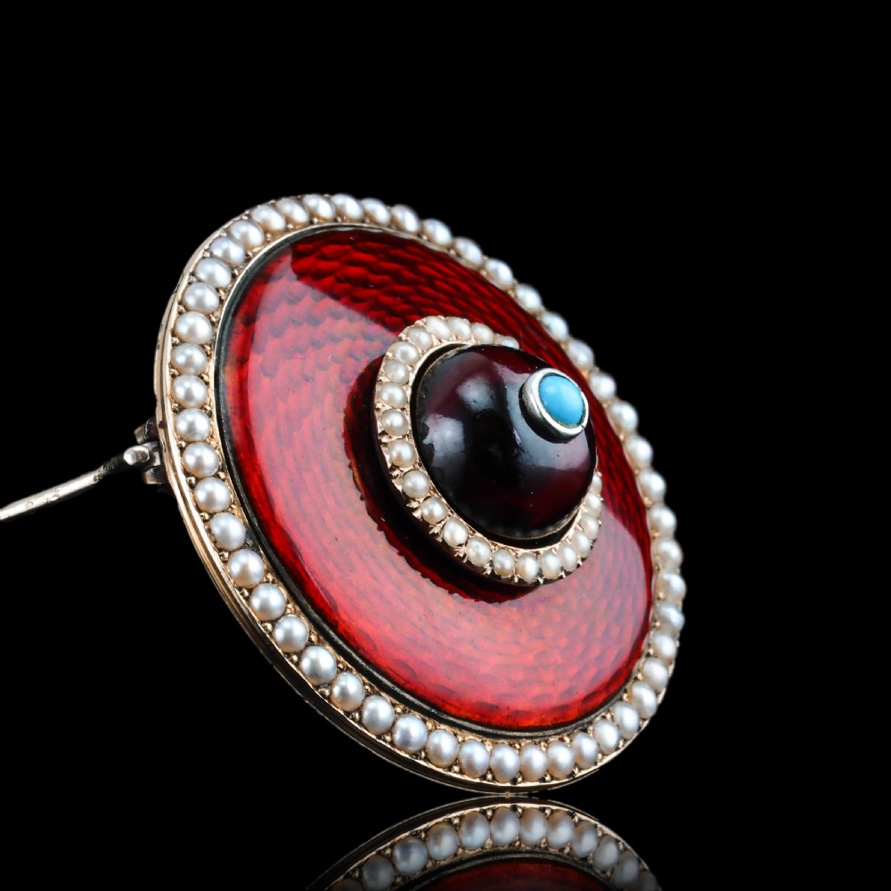 A circular Victorian red guilloche enamel brooch featuring concentric seed pearl borders, a central dark garnet cabochon, and a small turquoise accent set in gold. View III