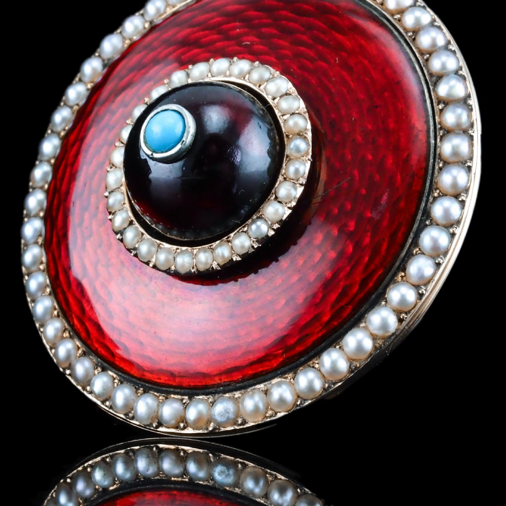 A circular Victorian red guilloche enamel brooch featuring concentric seed pearl borders, a central dark garnet cabochon, and a small turquoise accent set in gold. View IV